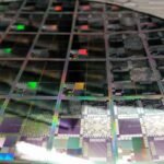 A close-up photograph of a large round semiconductor wafer containing an array of multicoloured microelectronic testbed dies, showing iridescent circuit patterns in purple, green, black and silver under bright lighting