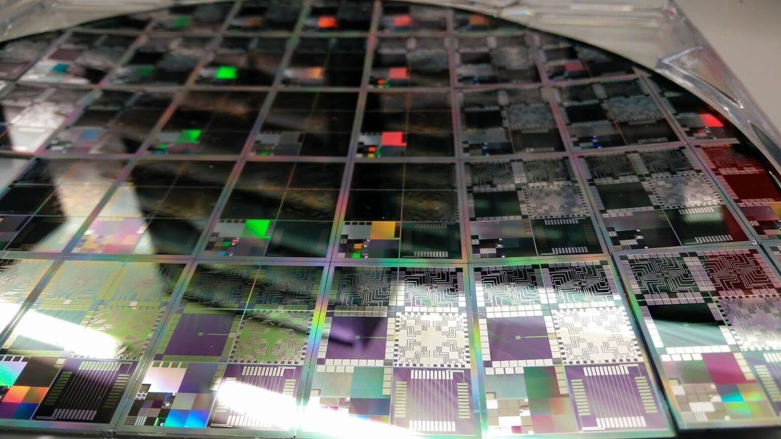 A close-up photograph of a large round semiconductor wafer containing an array of multicoloured microelectronic testbed dies, showing iridescent circuit patterns in purple, green, black and silver under bright lighting