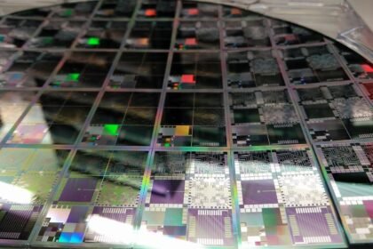 A close-up photograph of a large round semiconductor wafer containing an array of multicoloured microelectronic testbed dies, showing iridescent circuit patterns in purple, green, black and silver under bright lighting