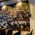 A large wall-mounted photographic mural inside a stock exchange building showing a busy trading floor filled with traders in white shirts and business attire, with electronic ticker boards visible at the top, surrounded by empty chairs and desks in the foreground
