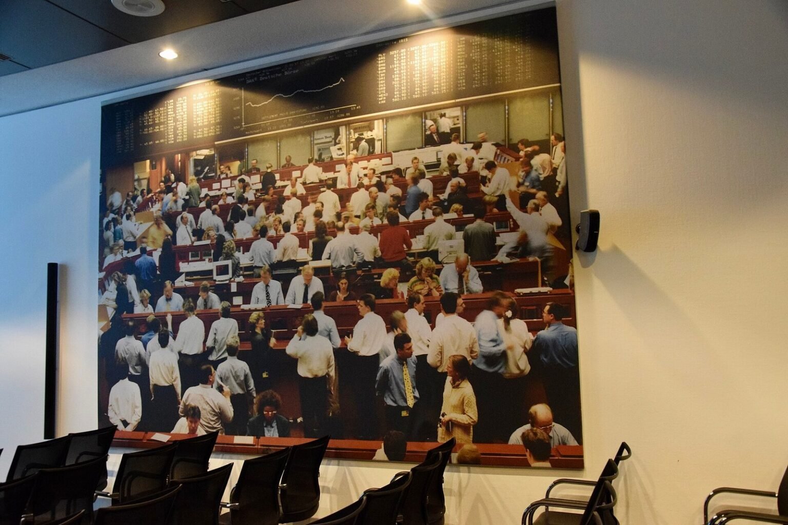A large wall-mounted photographic mural inside a stock exchange building showing a busy trading floor filled with traders in white shirts and business attire, with electronic ticker boards visible at the top, surrounded by empty chairs and desks in the foreground
